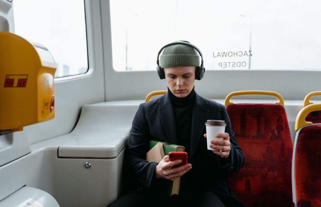Zdjęcie przedstawia mężczyznę w zielonej czapce, który słucha muzyki, trzyma w jednej dłoni kawę, a w drugiej czytnik do książek elektronicznych, jadąc autobusem.